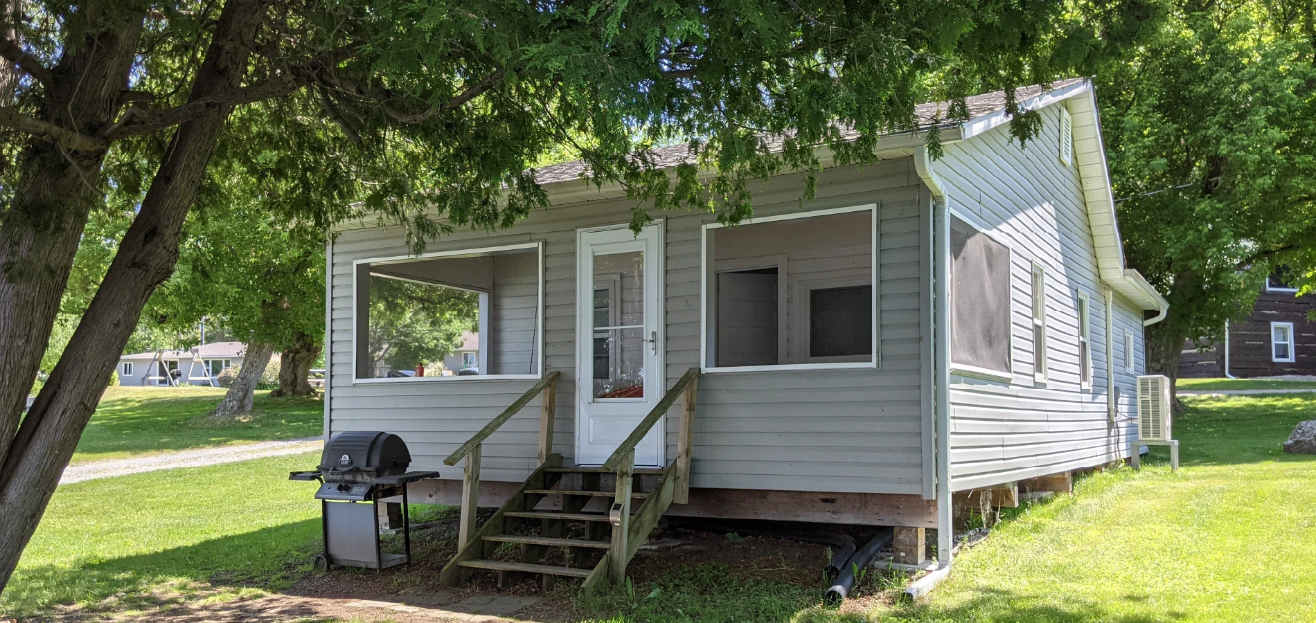 Pigeon Lake Resort Cottage five exterior with small porch, steps, barbecue, and trees providing shade.